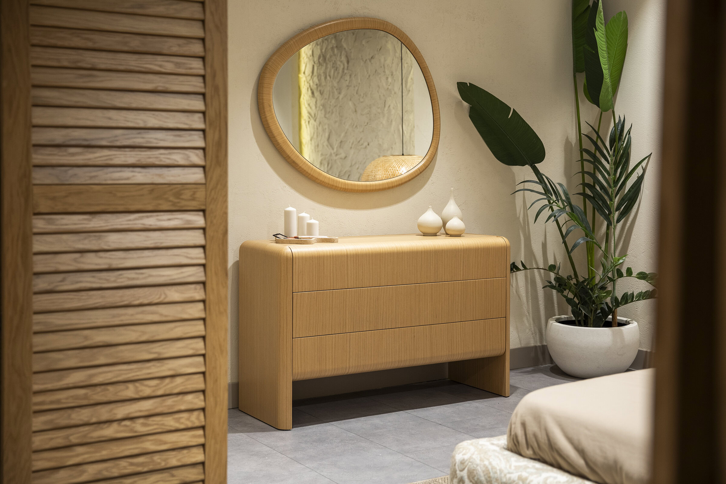 Natural wood dresser with sleek modern design, topped with decorative vases and candles, reflected in a stylish round mirror, accentuated by lush indoor plant in a contemporary white pot in a cozy home interior.