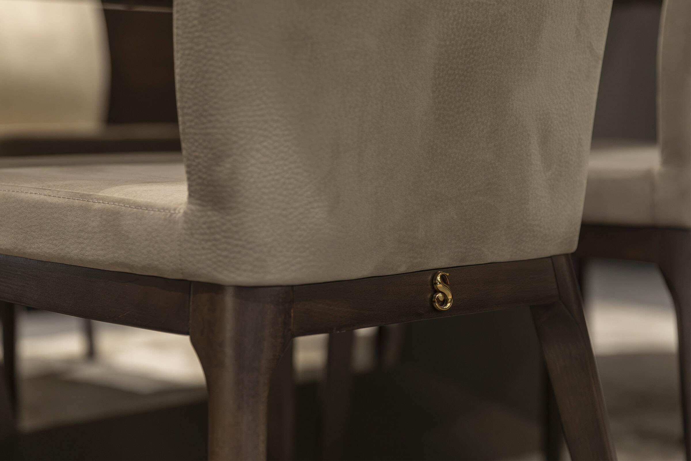 Luxurious beige upholstered dining chair with dark wooden legs and elegant gold S-shaped accent on the side. Perfect for sophisticated home interiors.