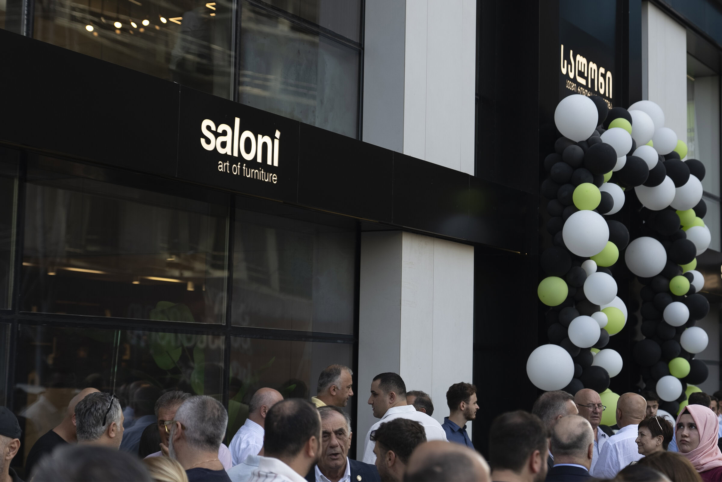 Saloni Furniture storefront with black exterior, festive black, white, and green balloon decoration, and a crowd of people outside.