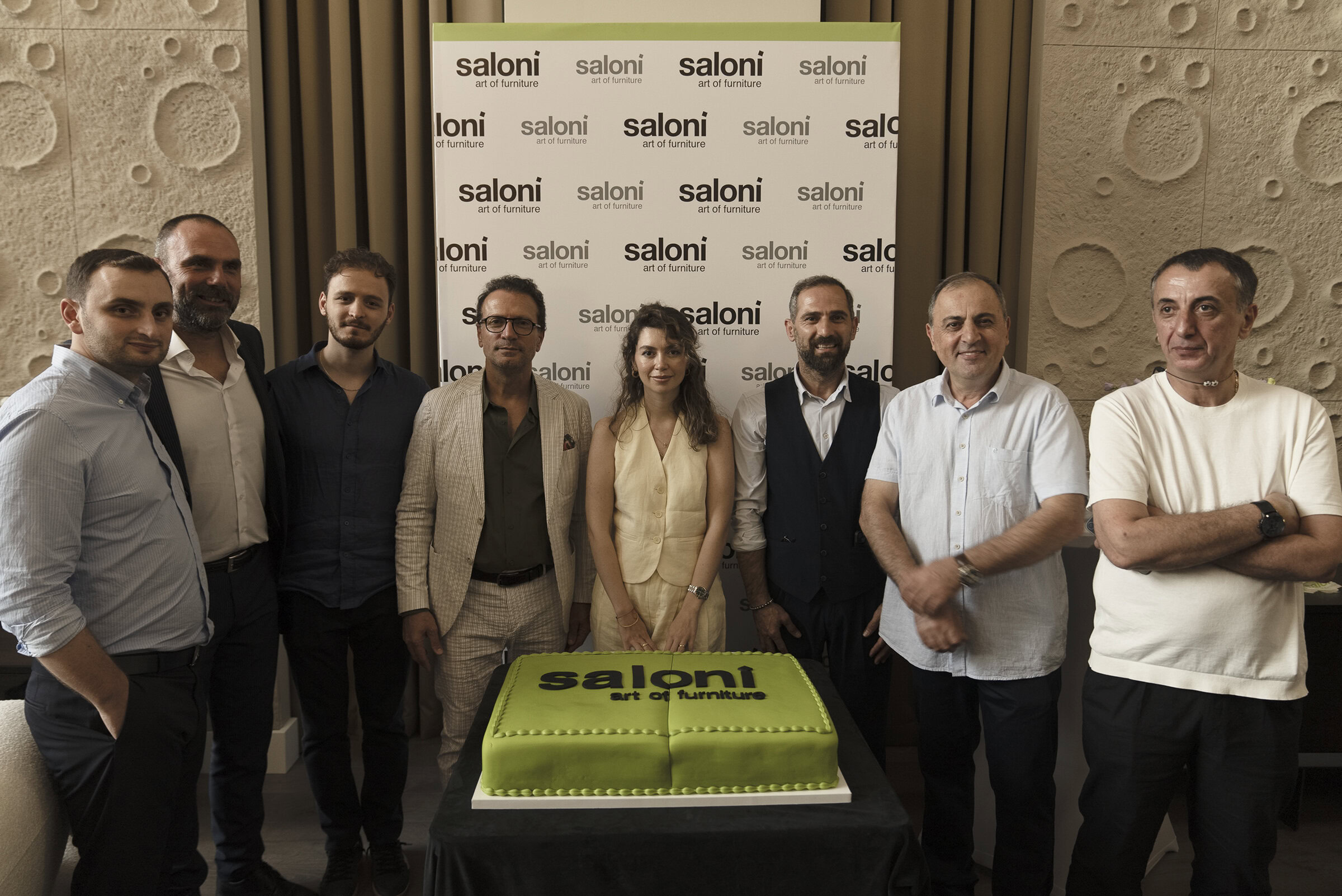 Elegant team of furniture designers celebrating Saloni Furniture event with a large green cake, showcasing the company's commitment to innovative furniture art and craftsmanship.